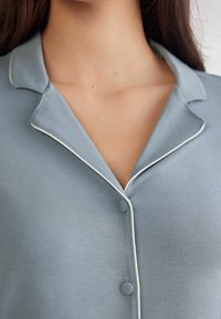 Světle modrá noční košile s límcem, bílou paspulí podél okrajů a kulatými knoflíky u výstřihu. Hladká textura a minimalistický design.