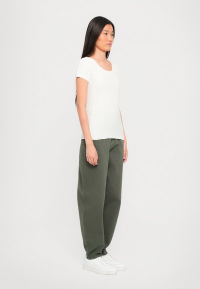 Witte, textuurige top met korte mouwen, gecombineerd met olijfgroene, losse broek en witte sneakers. Model staat in profiel tegen een lichte achtergrond.