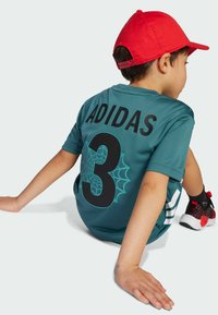 Maglietta Adidas color teal con la scritta nera "ADIDAS" e il numero "3," grafica a ragnatela. Maniche corte, texture del tessuto, abbinata a scarpe nere e rosse e un cappellino rosso.