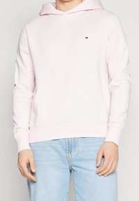 Sudadera con capucha de color rosa claro, hecha de un material suave, con un bolsillo delantero y un pequeño logo en la parte izquierda del pecho.