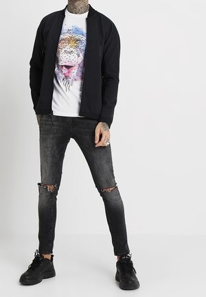 Person in schwarzer Jacke und weißem Grafik-T-Shirt mit buntem abstraktem Gesicht, in zerrissenen schwarzen Skinny-Jeans und schwarzen Sneakers, stehend vor einem weißen Hintergrund.