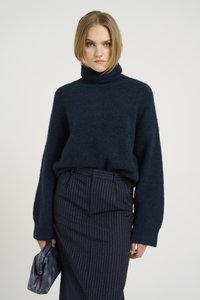 Granatowy sweter z golfem o teksturowanym splocie, zestawiony z ciemną spódnicą w prążki. Modelka trzyma małą, plisowaną szaro-skalistą torebkę.
