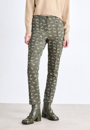 Vrouw draagt olijfgroene broek met zebraprint, beige trui en bijpassende olijfgroene laarzen met hak en kettinkdetail.