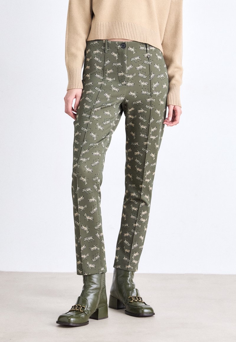 Femme portant un pantalon vert olive à imprimé zèbre, un pull beige et des bottines vert olive à talons avec un détail chaîne assorti.