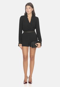 Blusa nera avvolgente con maniche lunghe, abbinata a pantaloni corti a vita alta. Presenta una cintura in catena dorata e una pochette nera, con tacchi a punta bordeaux.