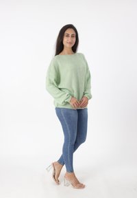 Grüner Strickpullover mit einem strukturierten Muster, weitem Ausschnitt und überschnittenen Schultern, kombiniert mit blauen Skinny Jeans und klaren Sandalen mit Absatz.