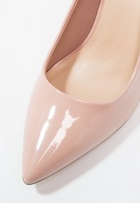 Chaussure à talon haut pointu rose clair brillant en gros plan montrant une texture lisse et une doublure intérieure beige sur fond blanc.