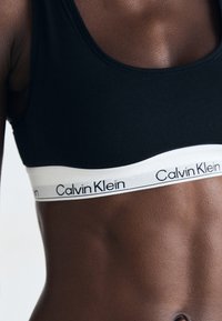 Svart sportsbh med bred hvit kant som har "Calvin Klein" merke. Glatt tekstur og tettsittende design.