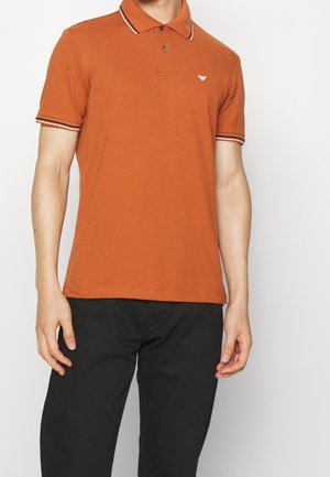 Pomarańczowa koszulka polo z kontrastującym paskowanym kołnierzykiem i mankietami na rękawach, krótkie rękawy, małe logo na piersi. Noszona z czarnymi spodniami.