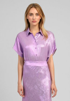 Femme portant un chemisier brillant lavande à manches courtes boutonné, rentré dans une jupe taille haute assortie, debout devant un fond uni.