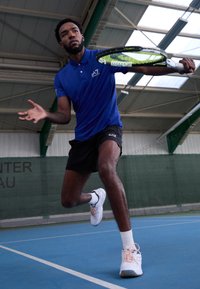 Idrettsutøver i blå polo-skjorte og svarte shorts, som forbereder seg på å treffe en tennisball med et grønt og svart racket på en blå bane.