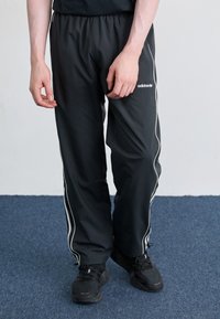Pantaloni sportivi negri cu dungi albe pe lateral, talie elastică și material ușor. Se poartă cu adidași negri și cu podeaua gri parțial vizibilă.