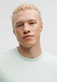 Gaiši zaļš T-krekls, kas izgatavots no mīksta materiāla, ar apaļu kakla izgriezumu un īsām piedurknēm, attēlots uz cilvēka ar gaišiem cirtainiem matiem.