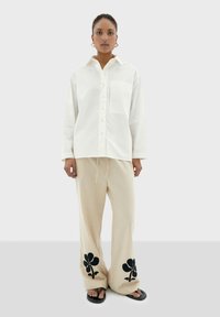 Camicia bianca con bottoni e tasca sul petto, abbinata a pantaloni larghi beige con ricami floreali neri all'orlo. Ciabatte nere.