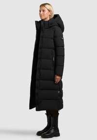Longue doudoune noire avec capuche, dotée d'une fermeture éclair, de poches latérales et de sections matelassées. Associée à des bottines noires.