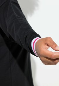 Musta pitkähihainen paita, jossa on teksturoitu kuvio ja valko- ja vaaleanpunaraidalliset kalvosimet. Käsi ojennettuna, näyttää luonnollisen ihon tekstuurin.