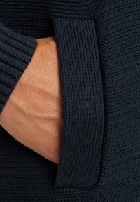 Donkerblauwe gebreide trui met een gestructureerde afwerking, voorzien van geribde mouwen en een zak, met een hand in de zak.