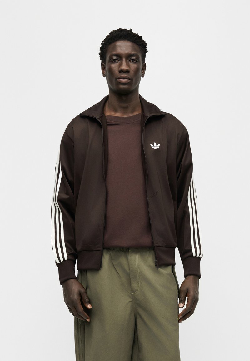 Uomo che indossa una giacca sportiva Adidas marrone scuro con strisce bianche sulle maniche, maglietta marrone e pantaloni verde oliva, in piedi contro uno sfondo bianco.