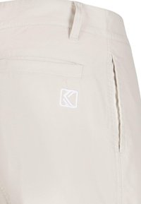 Šviesiai smėlio spalvos kelnės, su lygiu tekstūra, turi galinį kišenę ir mažą baltą logotipą. Matomas šoninis užtrauktukas.