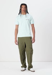 Lyseblå t-skjorte med rund hals og en liten logo på brystet, parret med løst sittende olivengrønne cargo-bukser med sidelommer. Hvite sneakers.