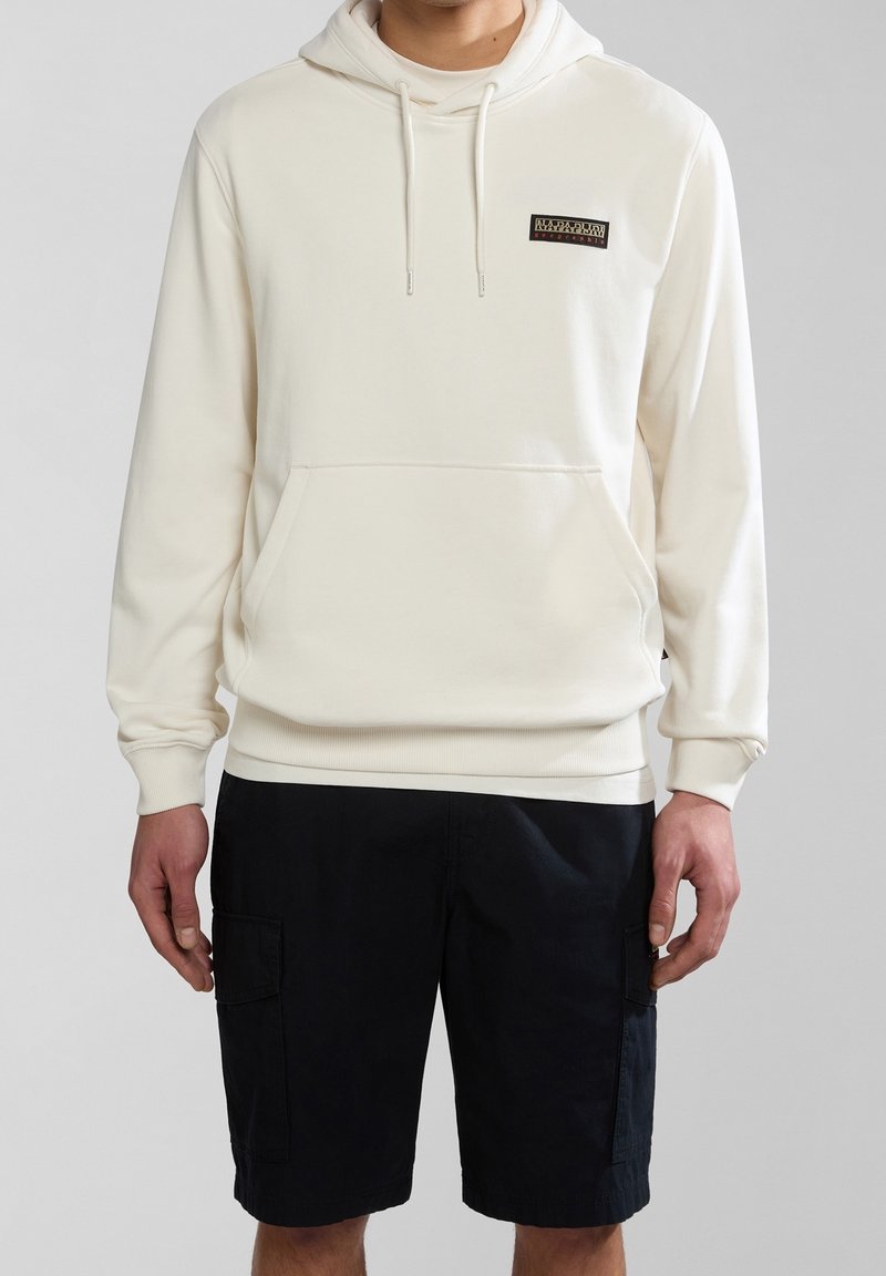 Personne portant un sweat à capuche crème avec un petit logo sur la poitrine et un short cargo noir, debout devant un fond clair et uni.