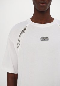 T-shirt en coton blanc à manches courtes, avec des accents en noir à motifs sur les épaules et un écusson rectangulaire sur la poitrine.