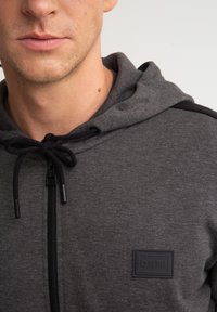 Szara bluza z kapturem zapinana na zamek, z czarnym akcentem na ramionach, z regulowanym kapturem oraz gumową metką na piersi. Gładka tekstura, casualowy design.