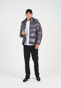 Grijs pufferjack met glanzende afwerking, ritszakken en afneembare capuchon, gecombineerd met een wit T-shirt en zwarte broek. Zwarte pet en sneakers.