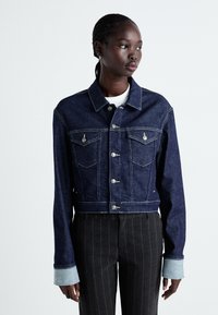 Mörkblå jeansjacka med en kroppsdesign, utrustad med två bröstfickor, silverknappar och kontrasterande ljusblå ärmar.