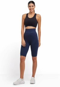 Reggiseno sportivo nero e pantaloni bike a vita alta blu navy con design a maglia testurizzata. Il modello indossa scarpe da ginnastica bianche e si trova in uno spazio neutro.