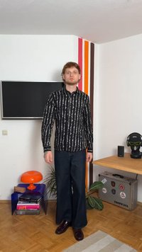 Schwarz-weiße gemusterte Hemd mit schlanker Passform, kombiniert mit weiten, dunklen Jeans und glänzenden schwarzen Schuhen. Innenraum mit dekorativen Elementen.