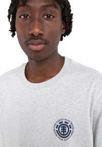 Graues Baumwoll-T-Shirt mit rundem Ausschnitt, das ein dunkelblaues, rundes Logo auf der linken Brust mit einer Baumgrafik und Elementwörtern aufweist.