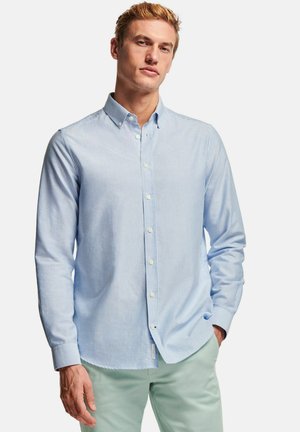 Man met een lichtblauw-wit gestreept overhemd en muntgroene broek, staand met één hand in de zak.