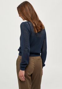 Mornarsko pleteno pulover z odprtim vzorcem, rebrastimi manšetami in robom. Zelo dobro se ujema s rjavimi hlačami, ki se ponašajo z ohlapnim krojem in gladko teksturo.