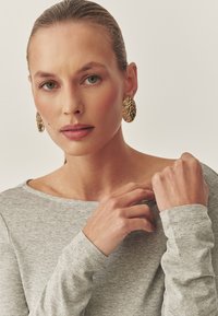 Grauer Langarmoberteil mit weitem Halsausschnitt; Modell trägt goldene strukturierte ovale Ohrringe und passt sanft den Kragen des Shirts an.