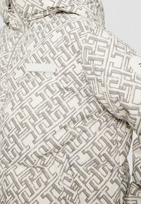 Doudoune blanche avec motif géométrique noir en forme de labyrinthe, fermeture éclair à l'avant et écusson circulaire sur la manche supérieure.