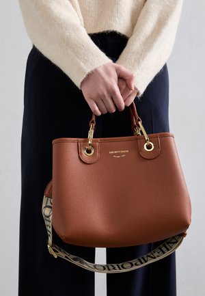 Mains tenant un sac à main en cuir marron Emporio Armani avec des ferrures dorées et une bandoulière en tissu de marque, porté avec un pull crème et un pantalon noir.