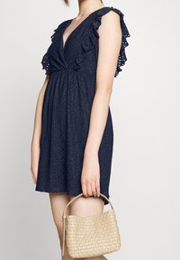 Sininen lyhyt mekko, jossa on frilla-hihat ja reikäpintakuvio, yhdistettynä kudottuun beigeen käsilaukkuun.