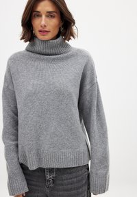 Gray turtleneck sweater with a relaxed fit, ribbed collar and hem, soft texture, and dropped shoulders. Worn with gray distressed jeans.