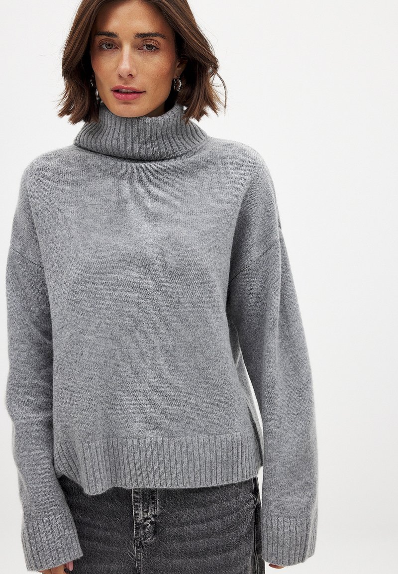 Suéter de cuello alto gris con un corte relajado, cuello y dobladillo de canalé, textura suave y hombros caídos. Combinado con jeans grises desgastados.