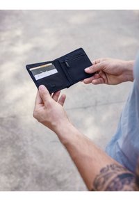 Black wallet shown open, featuring multiple card slots, a zippered pocket, and a smooth texture. Held in hands against a neutral background.