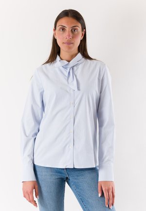 Donna con capelli castani lunghi che indossa una camicia a righe azzurro chiaro con cravatta al collo e jeans blu, in piedi davanti a uno sfondo bianco.