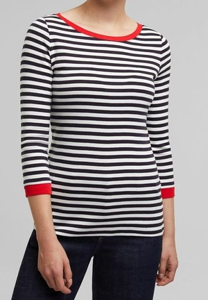 Personne portant un t-shirt rayé horizontal noir et blanc avec un col et des poignets rouges, associé à un jean foncé, debout devant un fond uni.