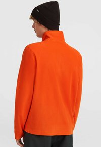 Person iført en lys orange fleecejakke og sort beanie, vendt væk mod en neutral lys baggrund.