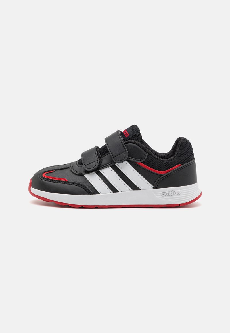 Zapatilla deportiva negra con dos correas ajustables de Velcro, rayas laterales blancas, acentos rojos y suela de goma. Presenta una parte superior texturizada para brindar soporte.