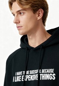 Sweat à capuche noir avec texte blanc : "JE DOIS RÉUSSIR PARCE QUE J'AIME LES CHOSES CHÈRES." Tissu doux et design décontracté.