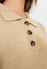 Beżowy sweter dzianinowy z kołnierzykiem, trzema ciemnymi okrągłymi guzikami, teksturowanym materiałem i widokiem z bliska na ramiona.