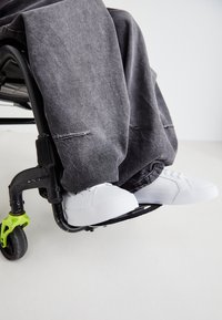 Bele teniske na naslonu za noge invalidskih kolica, u kombinaciji s tamnosivim širokim hlačama. Invalidska kolica imaju crni okvir i zeleni naglasak na točkovima.