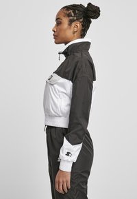 Veste coupe-vent noir et blanc avec un col haut, une taille élastique et un accent logo sur la poitrine. Tissu lisse et léger.