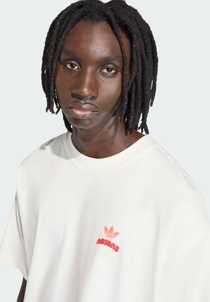 Junge Person mit mittel-dunkler Haut und schulterlangen Dreadlocks trägt ein weißes Adidas-T-Shirt mit einem kleinen roten Logo auf der Brust.